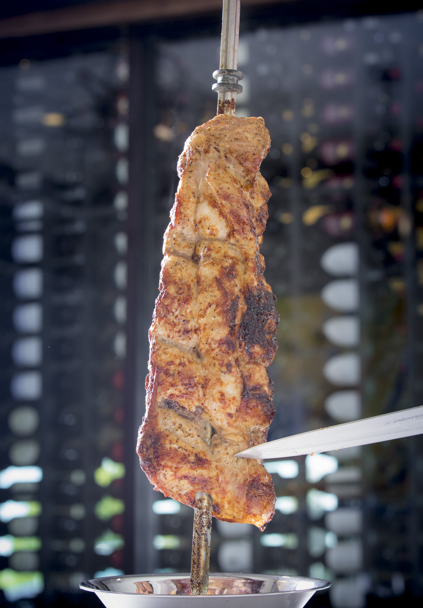 Grilled meat on skewer being carved tableside, showing caramelized exterior and juicy interior with carving knife and collection plate below.