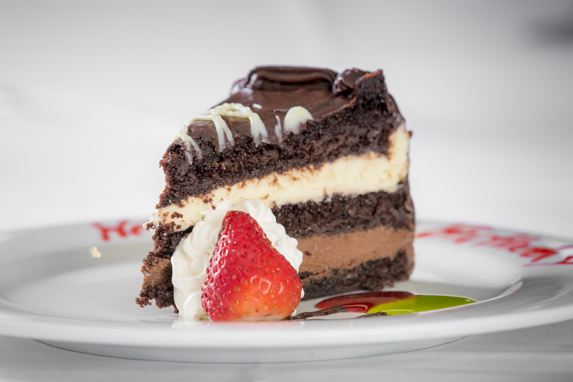 Chocolate layer cake slice with whipped cream, chocolate shavings, fresh strawberry, and mint garnish on white plate.