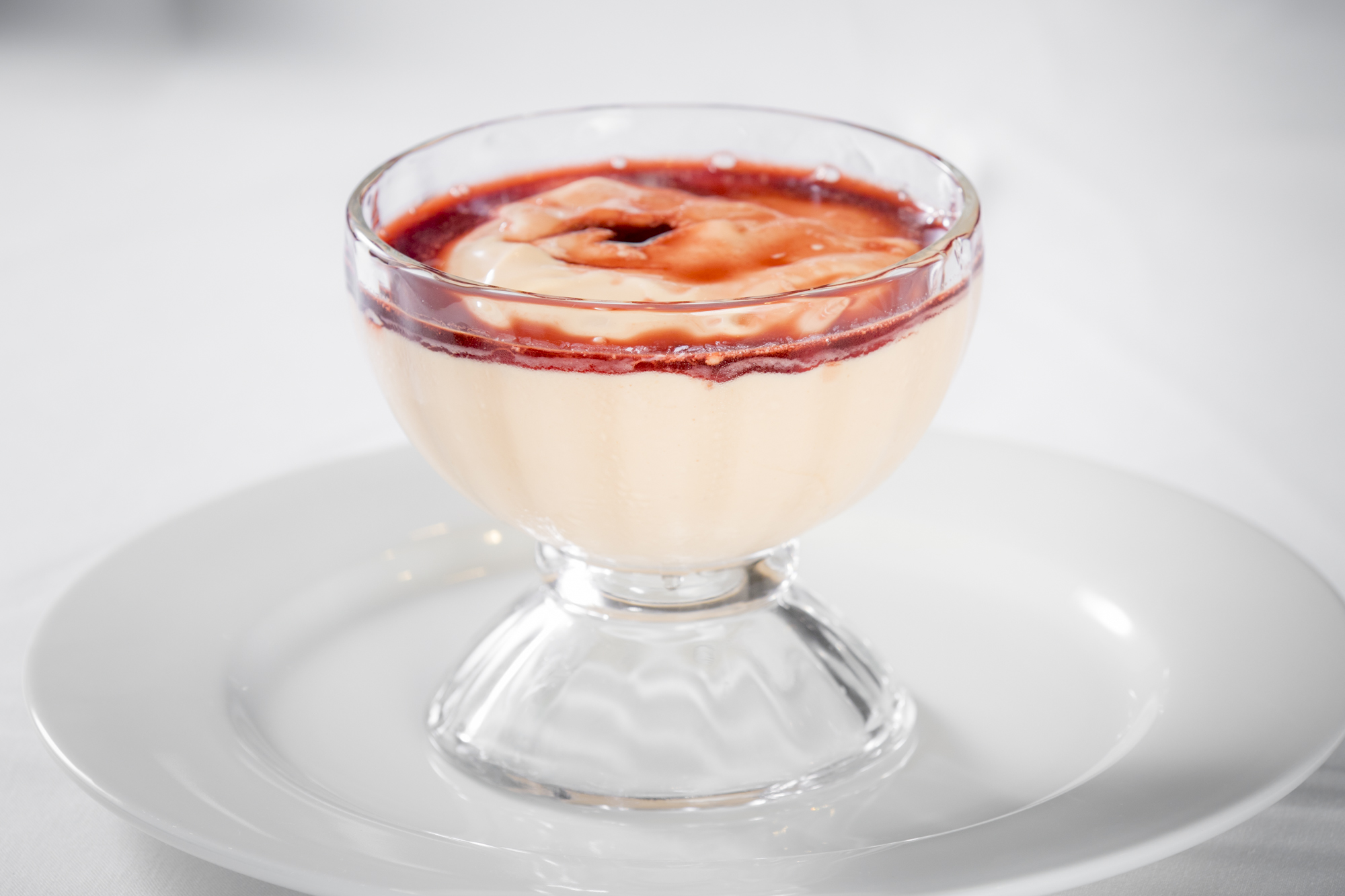 Layered dessert in a coupe glass with creamy custard base and red fruit coulis topping on a white plate.