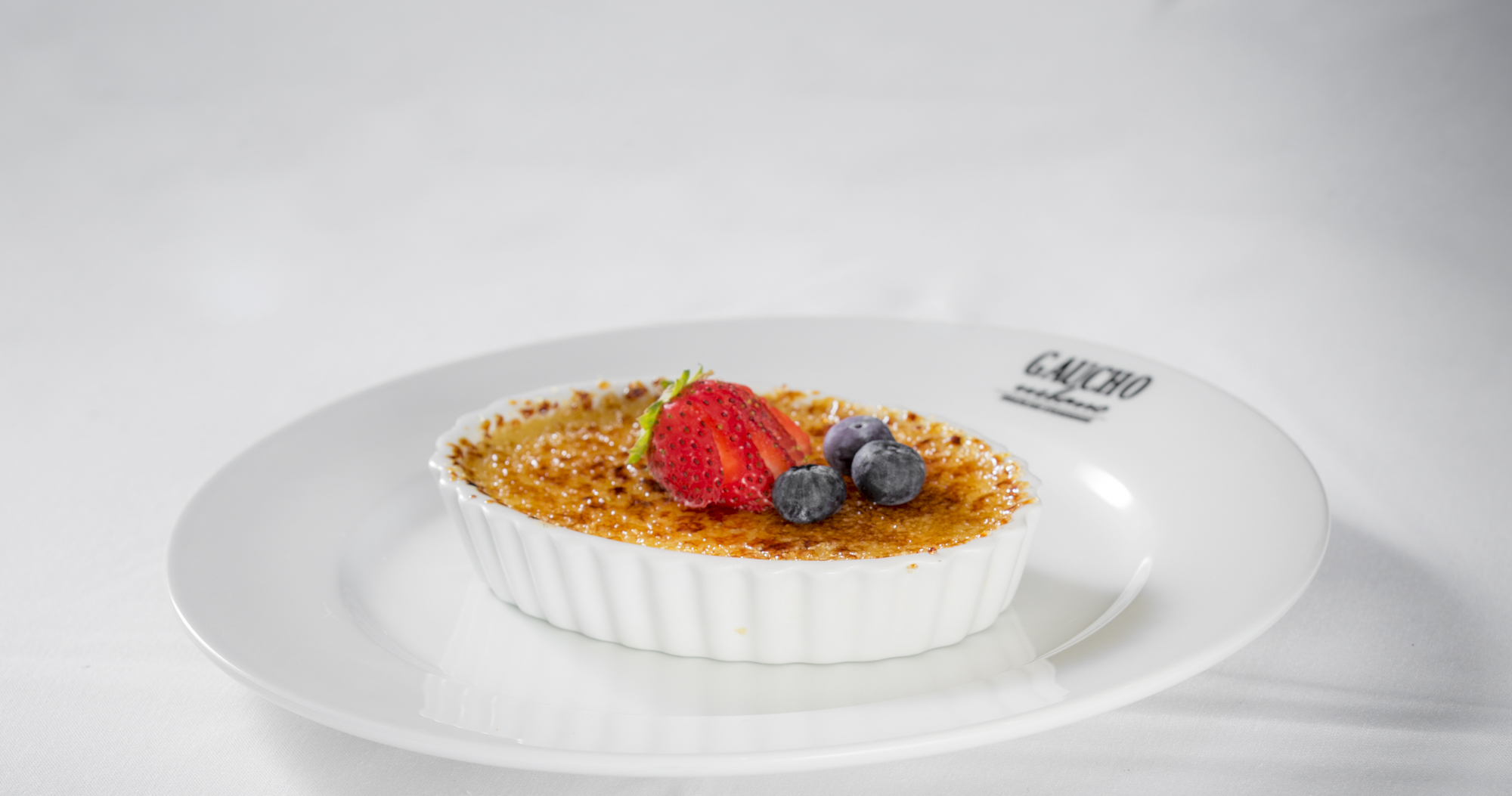 Crème brûlée dessert in white ramekin topped with caramelized sugar, fresh strawberry, blueberries, and mint leaf on white plate with restaurant logo.