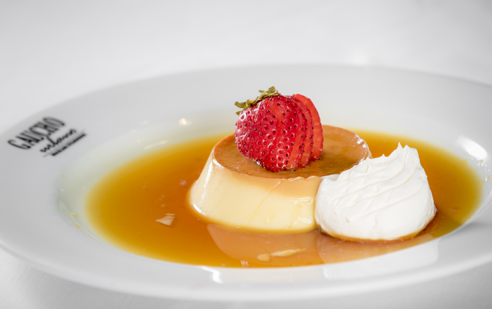 Flan dessert plated in caramel sauce with whipped cream and fresh strawberry garnish.